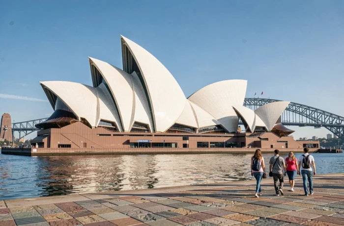 Vue panoramique de l'Opéra de Sydney depuis le port, avec le Harbour Bridge en arrière-plan et des touristes sur l'esplanade